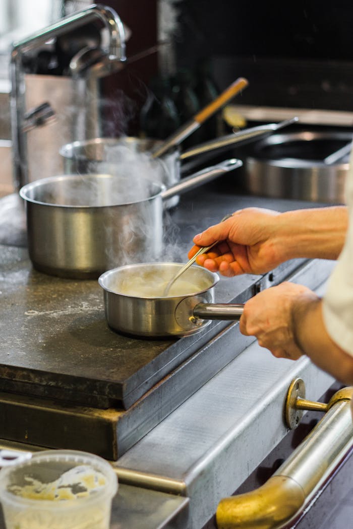 hero-img-01 A chef's hands stirring sauce in a stainless steel pan, creating steam in a professional kitchen setting.